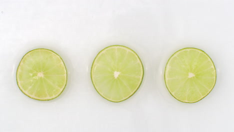 three-slices-of-lime-lie-on-white-in-the-background-in-slow-motion-falling-water-splashes.